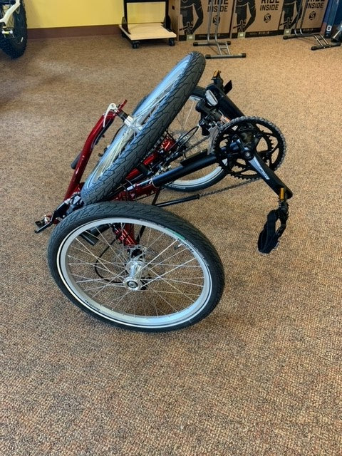 Folded view of a red and black TerraTrike Traveler recumbent trike with a black seat and folding handlebars, equipped with a chain drive and mechanical disc brakes.